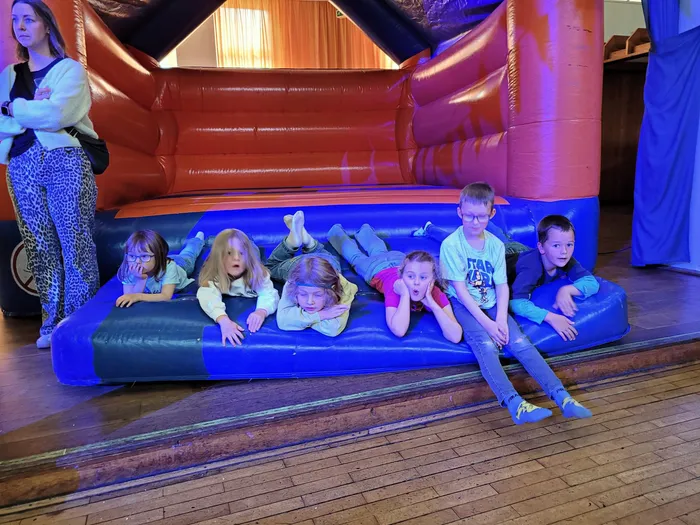 Sechs Kinder liegen und sitzen auf einer blauen Hüpfburg vor rotem Hintergrund. Im Hintergrund steht eine Frau und beobachtet die Kinder.