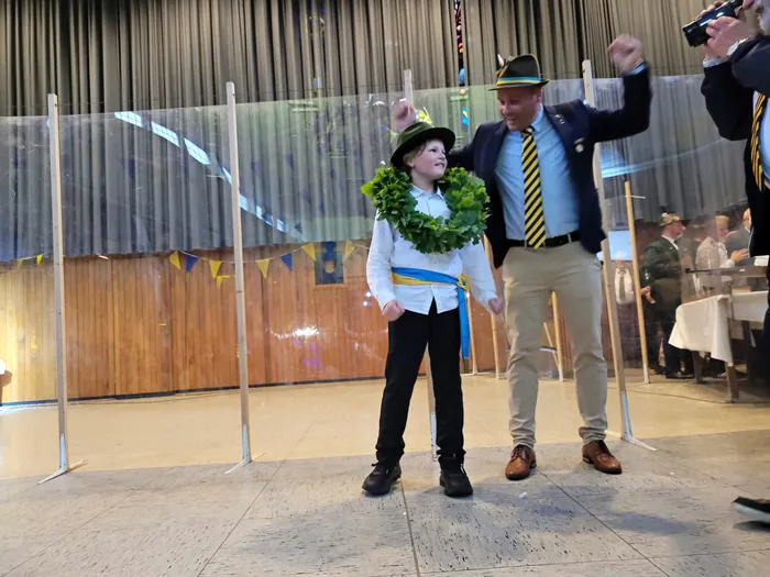 Ein Junge mit einem grünen Kranz um den Hals und ein Mann in Tracht gehen lächelnd durch einen Raum voller Menschen. Der Junge wurde zum Kinderschützenkönig ernannt.