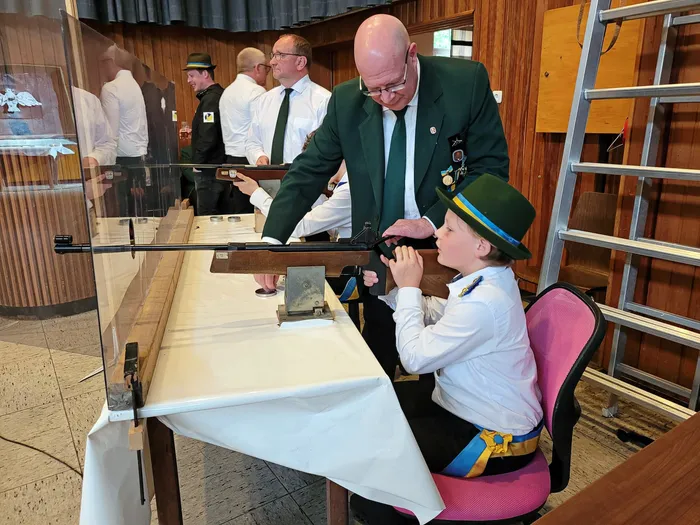 Ein Mann in grünem Jackett erklärt einem Jungen mit Schützenhut und Uniform die Handhabung eines Gewehrs. Im Hintergrund stehen weitere Männer in Schützenuniformen.