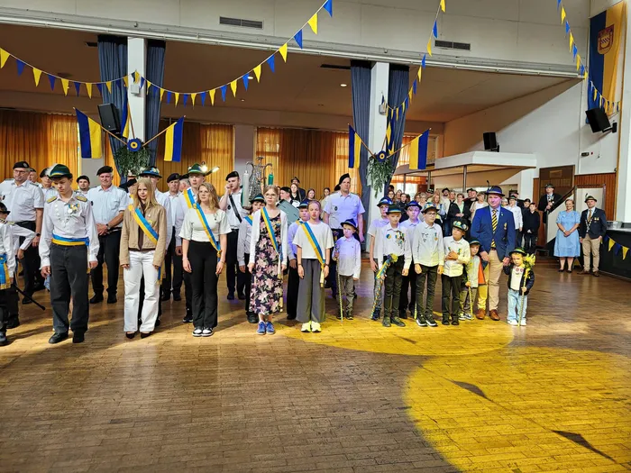 Eine Gruppe von Menschen, darunter Kinder und Erwachsene in Uniformen oder festlicher Kleidung, steht in einem hellen Raum mit Parkettboden. Viele tragen Bänder oder Abzeichen und blicken in die Kamera. Im Hintergrund sind blau-gelbe Girlanden zu sehen.