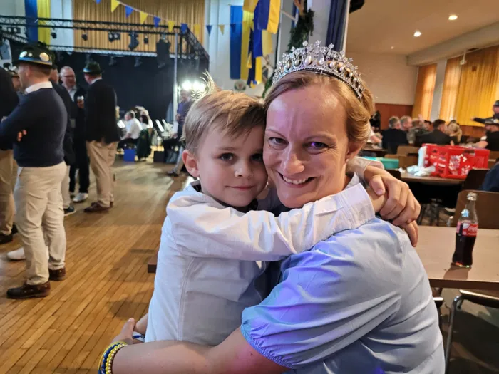 Eine Frau mit einer Tiara umarmt einen Jungen. Im Hintergrund sind Menschen in einem Festsaal zu sehen.
