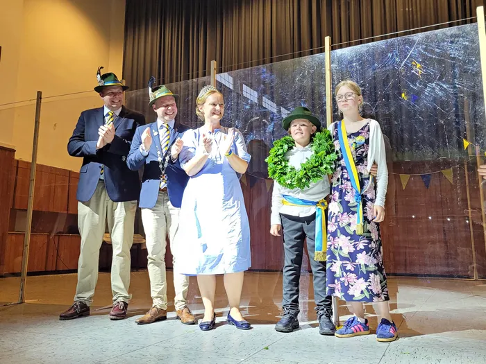 Fünf Personen stehen auf einer Bühne und applaudieren. Links ein Mann im Anzug mit Hut, daneben ein Mann mit Orden, in der Mitte eine Frau mit Krone, rechts ein Kind mit Blumenkranz und eine junge Frau mit Blumenkette. Im Hintergrund eine verschwommene Wand mit Girlanden.