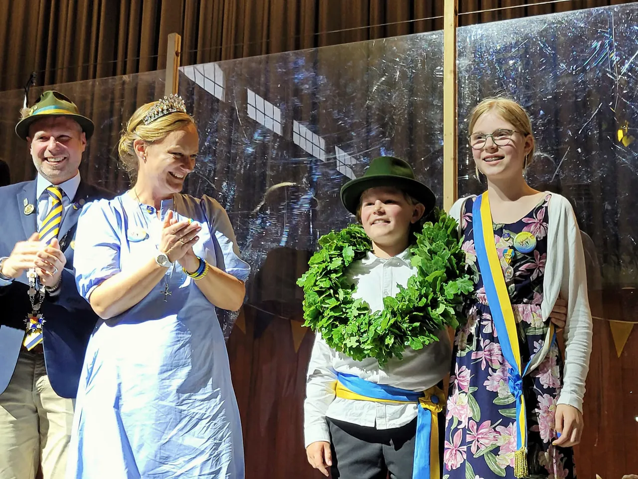Ein Mann, eine Frau mit Tiara, ein Junge und ein Mädchen stehen auf einer Bühne. Der Junge trägt einen grünen Hut und einen grünen Kranz, das Mädchen einen blumigen Schärpe und eine goldene Kette. Die Frau klatscht in die Hände, während der Mann lächelt. Im Hintergrund ist ein verschwommener Wald zu sehen.