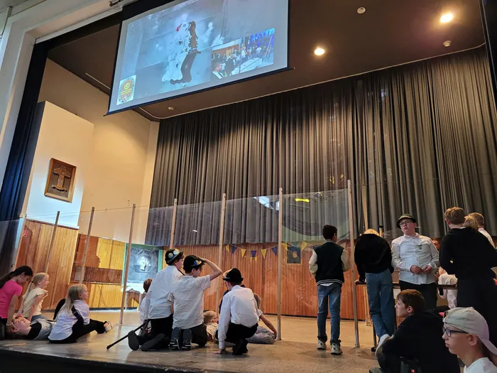 Kinder in historischen Kostümen sitzen und stehen auf einer Bühne vor einem Vorhang. Auf einer Leinwand im Hintergrund ist ein Bild von einer Gruppe von Menschen zu sehen.