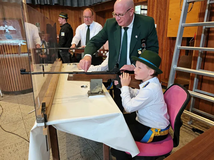 Ein Mann in grünem Jackett zeigt einem Jungen mit grüner Mütze und weißem Hemd, wie man mit einem Gewehr umgeht. Im Hintergrund stehen weitere Männer in Anzügen und Uniformen.