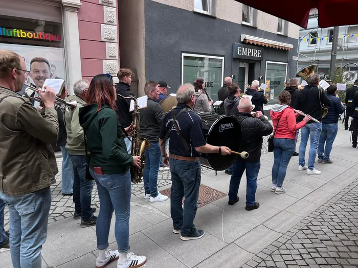 Eine Straßenkapelle spielt vor Geschäften in einer Fußgängerzone. Mehrere Musiker spielen Blechblasinstrumente und eine Trommel.