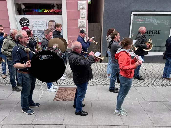 Eine Straßenkapelle spielt während des Schützenfestes der Plettenberger Schützengesellschaft. Mehrere Musiker mit Blechblasinstrumenten und einer Pauke stehen und spielen auf einer Straße vor Geschäften.