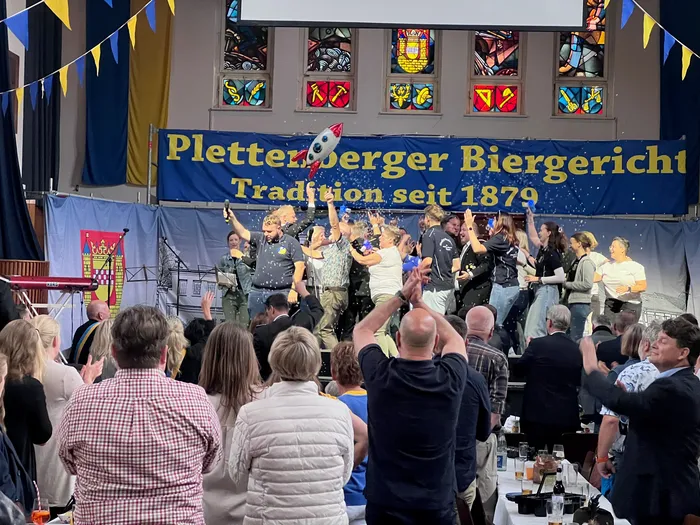 Eine Gruppe von Menschen feiert in einem Innenraum, Konfetti fliegt durch die Luft. Im Hintergrund ein Banner mit der Aufschrift „Plettenberger Biergericht, Tradition seit 1879“.