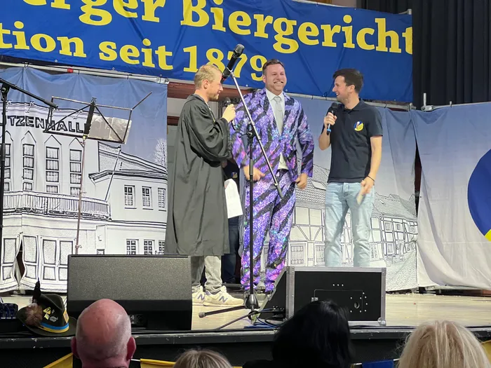 Drei Personen stehen auf einer Bühne vor einem Banner mit der Aufschrift „Plettenberger Biergericht seit 1879“. Ein Mann in einer Robe reicht einem Mann im karierten Gewand ein Zepter. Ein dritter Mann hält ein Mikrofon.