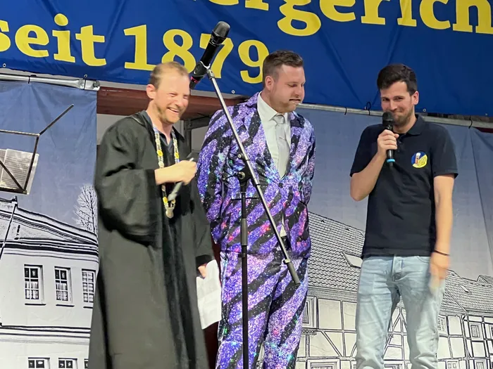 Drei Männer stehen auf einer Bühne vor einem Banner mit der Aufschrift „seit 1879“ und sprechen in Mikrofone. Der Mann in der Mitte trägt einen auffälligen, gemusterten Anzug.