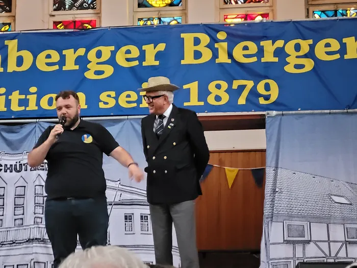 Zwei Männer stehen auf einer Bühne vor einem Banner mit der Aufschrift „Plettenberger Biergarten Tradition seit 1879“. Der Mann links hält ein Mikrofon, der Mann rechts trägt eine Uniform.