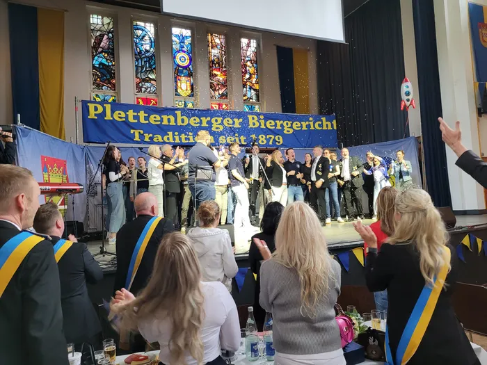 Eine Gruppe von Menschen steht auf einer Bühne vor einem Banner mit der Aufschrift „Plettenberger Biergericht, Tradition seit 1879“. Konfetti fällt herab, während das Publikum applaudiert.