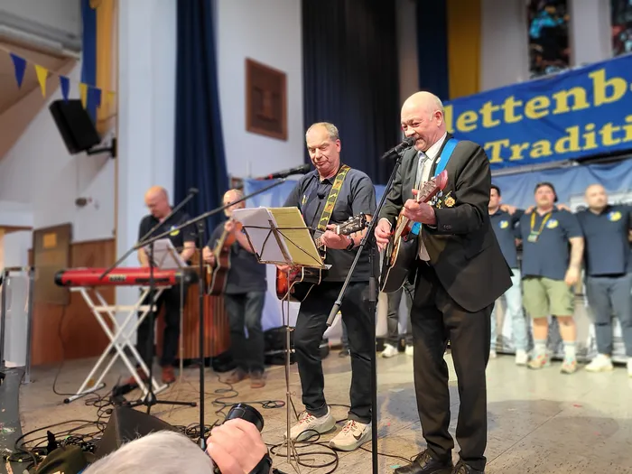 Zwei Musiker spielen Gitarre und singen in ein Mikrofon auf einer Bühne. Im Hintergrund sind weitere Musiker und ein Banner mit der Aufschrift „Plettenberger Schützenfest“ zu sehen.