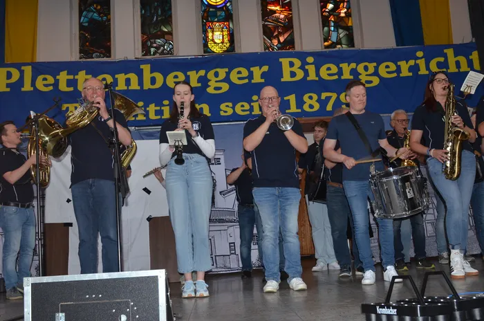 Eine Blaskapelle spielt auf einer Bühne vor einem Banner mit der Aufschrift „Plettenberger Biergericht – Tradition seit 1879“. Die Musiker spielen verschiedene Instrumente wie Posaune, Saxophon und Schlagzeug.