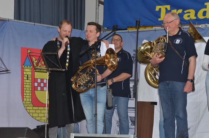 Vier Männer stehen auf einer Bühne und spielen Posaunen. Links hält einer ein Mikrofon, während die anderen drei Blasinstrumente spielen. Im Hintergrund ist ein Banner mit dem Wappen der Stadt Plettenberg zu sehen.