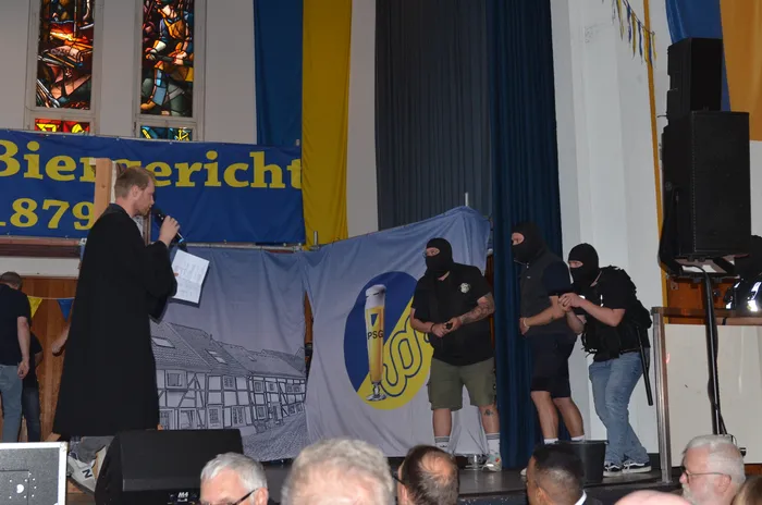Ein Mann in schwarzer Robe hält ein Mikrofon vor einer Gruppe maskierter Personen, die aneinandergekettet sind. Im Hintergrund ein Banner mit der Aufschrift 