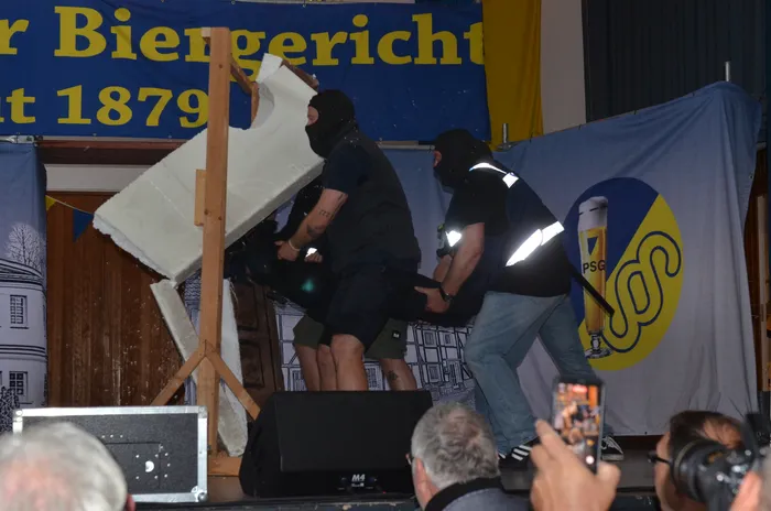 Zwei Personen in Arbeitskleidung zerreißen gemeinsam ein großes Stück Papier auf einer Bühne vor einem Banner mit der Aufschrift 