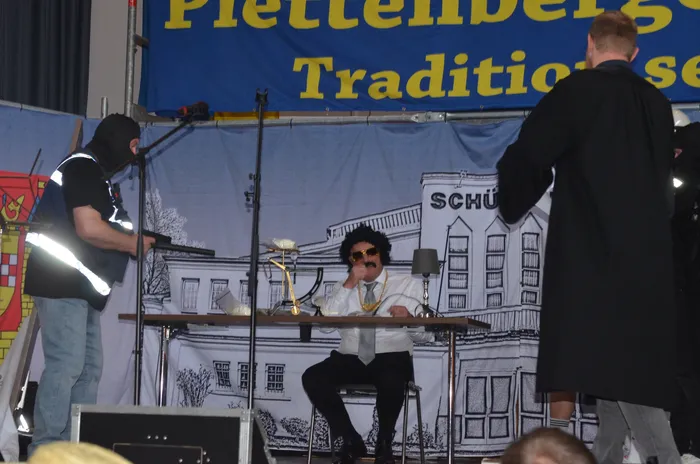 Ein Mann mit dunkler Perücke und Sonnenbrille sitzt an einem Tisch mit Getränken und Snacks. Links steht ein Mann in Montur und hält ein Mikrofon, während im Hintergrund ein Banner mit der Aufschrift 