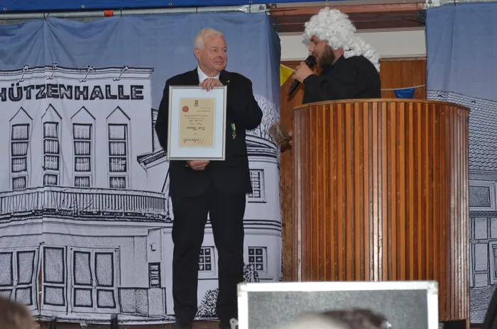 Ein Mann in dunklem Anzug hält eine Urkunde mit einem Wappen und Text entgegen, während im Hintergrund ein Mann mit weißer Perücke am Rednerpult steht. Im Hintergrund ist ein Banner mit der Aufschrift „Schützenhalle“ zu sehen.