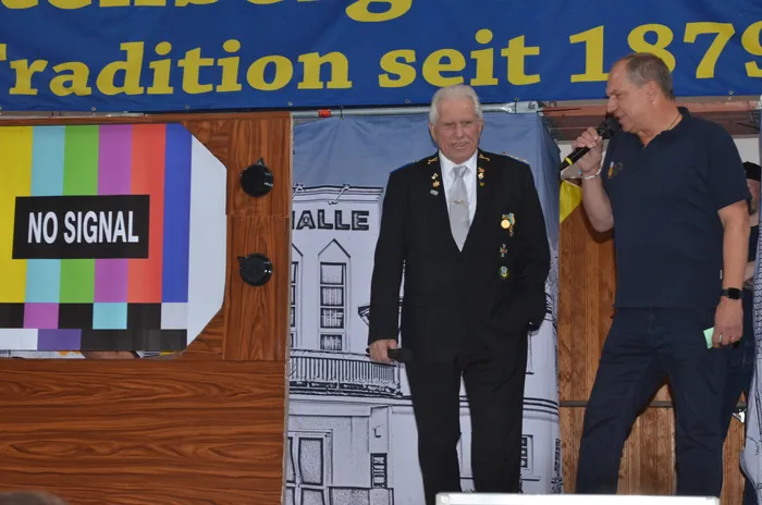 Ein Mann in einem dunklen Anzug steht auf einer Bühne und hält ein Mikrofon. Neben ihm steht ein weiterer Mann, der ebenfalls ein Mikrofon hält. Im Hintergrund ein Banner mit der Aufschrift „Tradition seit 1879“ und ein Schild mit der Aufschrift „No Signal“.