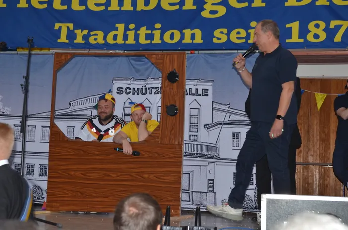 Ein Mann in einem Deutschlandtrikot sitzt hinter einem Pult mit Mikrofonen, während ein zweiter Mann mit Mikrofon auf einer Bühne steht. Im Hintergrund ein Banner mit der Aufschrift „Plettenberger Schützenfest Tradition seit 1871“.