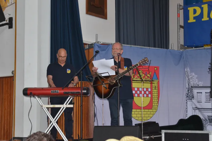 Zwei Männer auf einer Bühne, einer spielt Keyboard, der andere liest von einem Blatt ab und hält eine Gitarre. Im Hintergrund ein Banner mit einem Wappen und blauem Stoff.