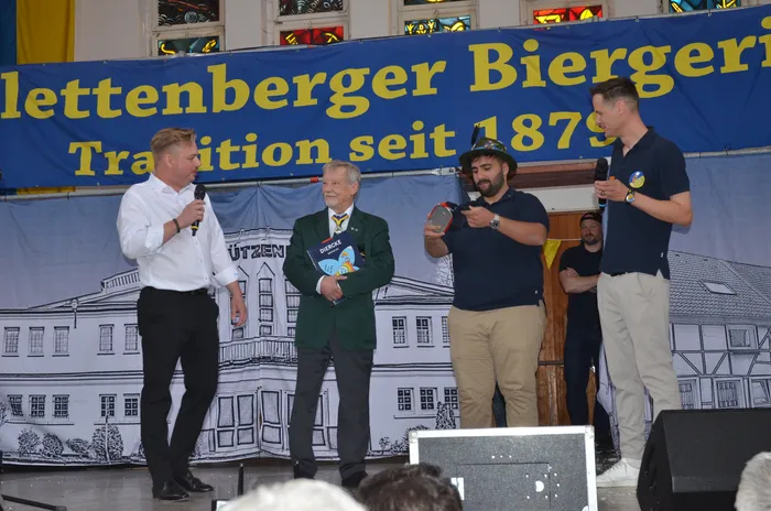 Vier Männer stehen auf einer Bühne vor einem Banner mit der Aufschrift „Plettenberger Biergericht, Tradition seit 1879“. Zwei der Männer halten Mikrofone, während der Mann in der Mitte eine Anstecknadel trägt. Im Hintergrund ist eine stilisierte Darstellung eines Gebäudes zu sehen.