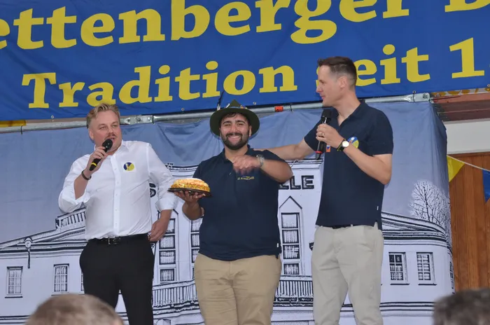 Drei Männer stehen auf einer Bühne, einer hält einen gefüllten Tablett mit Speisen. Im Hintergrund ein blaues Banner mit der Aufschrift 