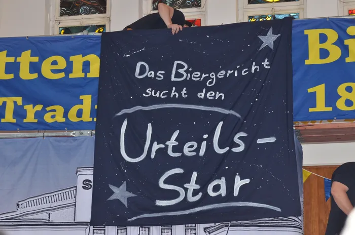Ein dunkler Banner mit weißer Schrift kündigt an: „Das Biergericht sucht den Urteils-Star“. Im Hintergrund sind weitere Banner und Teile von Zuschauern zu sehen.