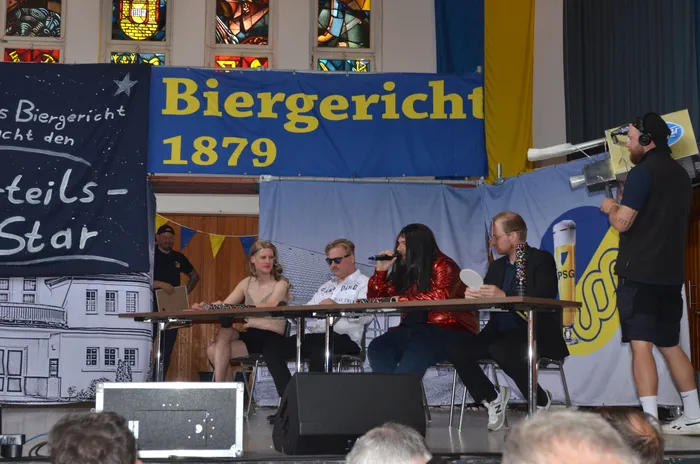 Fünf Personen sitzen an einem Tisch vor einem Banner mit der Aufschrift „Biergericht 1879“. Ein Mann singt in ein Mikrofon, während die anderen zuhören. Im Hintergrund ein Fenster mit Buntglas und ein Mann mit einer Kamera.