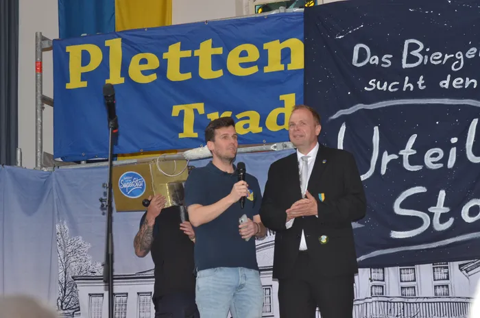 Zwei Männer mit Mikrofonen stehen vor einem Banner mit der Aufschrift „Plettenberg“ und „Das Biergericht sucht den SuperStar“. Im Hintergrund ist ein stilisiertes Stadtsilhouetten zu sehen.