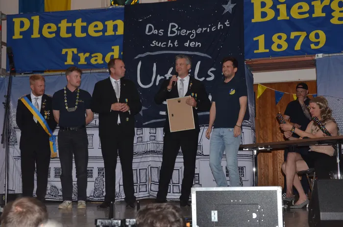 Fünf Männer stehen auf einer Bühne vor einem Banner mit der Aufschrift „Das Biergericht sucht den Urteil“. Der Mann in der Mitte hält eine braune Urkunde. Zwei Frauen sitzen rechts auf Stühlen.