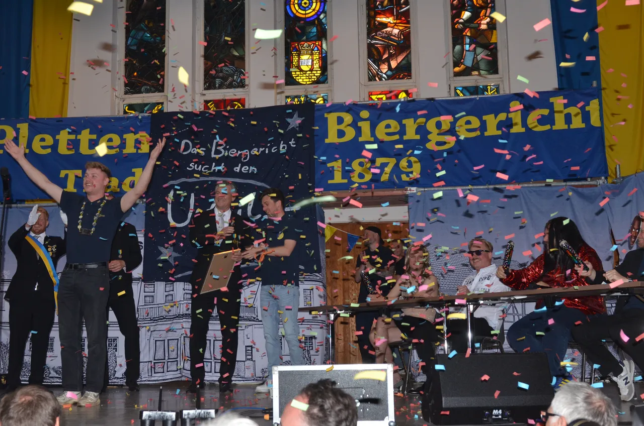 Eine Gruppe von Menschen steht auf einer Bühne vor einem Banner mit der Aufschrift „Biergericht 1879“. Konfetti fällt herab, während eine Person eine Auszeichnung entgegennimmt. Im Hintergrund sind Buntglasfenster zu sehen.