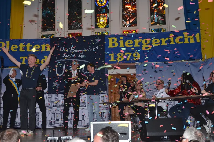Eine Gruppe von Menschen steht auf einer Bühne vor einem Banner mit der Aufschrift „Biergericht 1879“. Konfetti fällt herab, während eine Person eine Auszeichnung entgegennimmt. Im Hintergrund sind Buntglasfenster zu sehen.