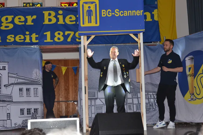 Ein Mann in einem dunklen Anzug mit vielen Orden steht auf einer kleinen Bühne mit erhobenen Armen. Über ihm hängt ein blaues Schild mit der Aufschrift „BG-Scanner“. Im Hintergrund ein Banner mit der Aufschrift „Berger Bier seit 1879“.