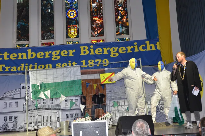 Ein Mann in dunkler Kleidung hält eine Rede vor zwei Personen in weißen Schutzanzügen und gelben Masken auf einer Bühne. Im Hintergrund ein Banner mit der Aufschrift 