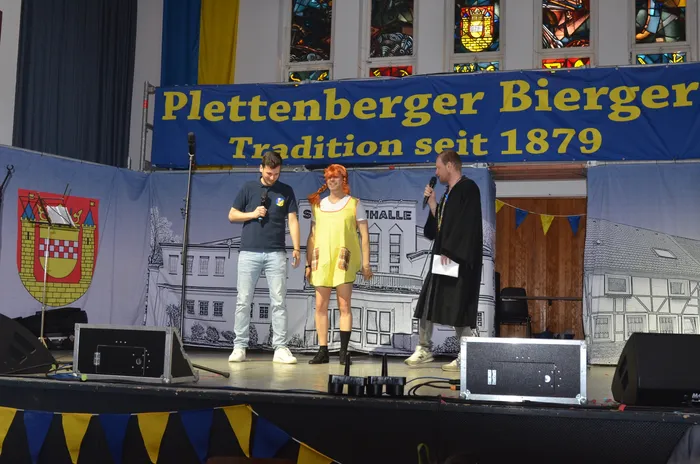 Zwei Männer und eine Frau stehen auf einer Bühne vor einem Banner mit der Aufschrift „Plettenberger Bierger Tradition seit 1879“. Im Hintergrund ist ein Wappen und eine stilisierte Stadtsilhouette zu sehen.