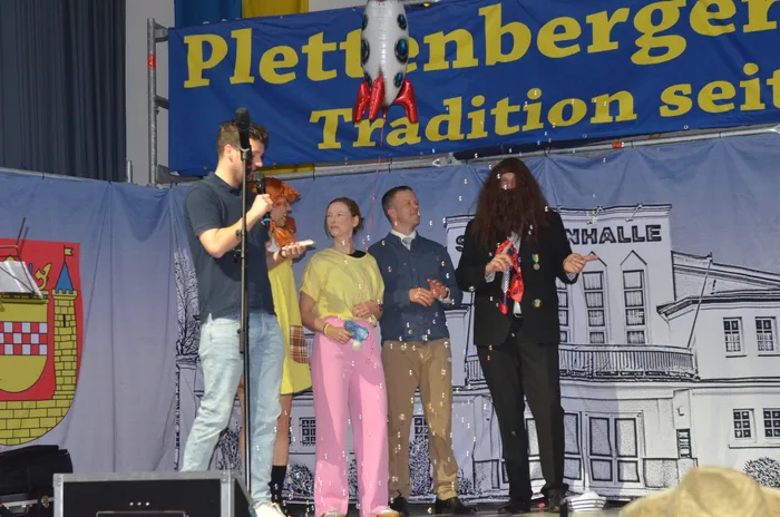 Vier Personen stehen auf einer Bühne vor einem Banner mit der Aufschrift „Plettenberger Tradition seit…“. Konfetti fällt herab. Im Hintergrund ist ein Gebäude mit der Aufschrift „Halle“ zu sehen.