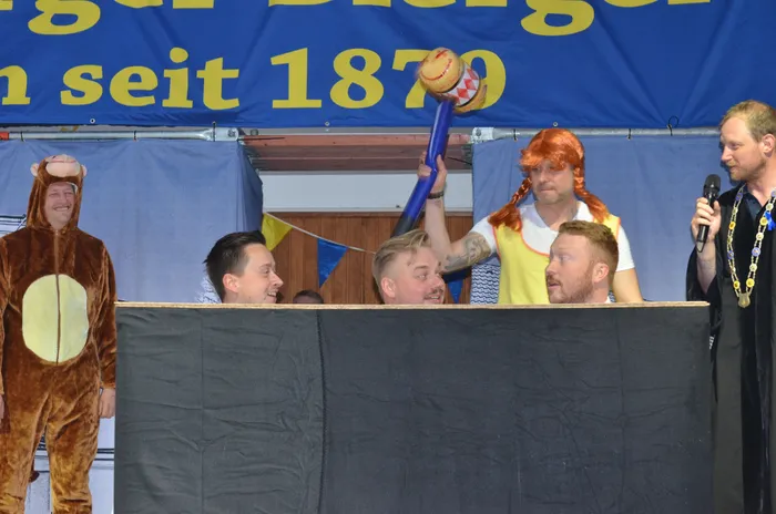 Fünf Männer stehen auf einer erhöhten Plattform. Links trägt einer einen Bärenkostüm, daneben stehen drei weitere Männer, einer davon mit rotem Haar und gelbem Hemd. Rechts hält ein Mann ein Mikrofon. Im Hintergrund ein blaues Banner mit der Aufschrift „seit 1879“.