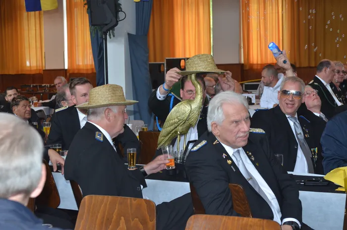 Männer in dunklen Anzügen und Strohhüten sitzen an einer langen Tafel mit Biergläsern und Speisen. Im Vordergrund hält eine Person eine goldene Kranichfigur. Im Hintergrund weitere Personen und eine blau-gelbe Flagge.