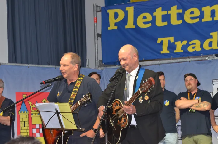 Zwei Männer spielen Gitarre und singen in ein Mikrofon auf einer Bühne. Im Hintergrund ein Banner mit der Aufschrift 