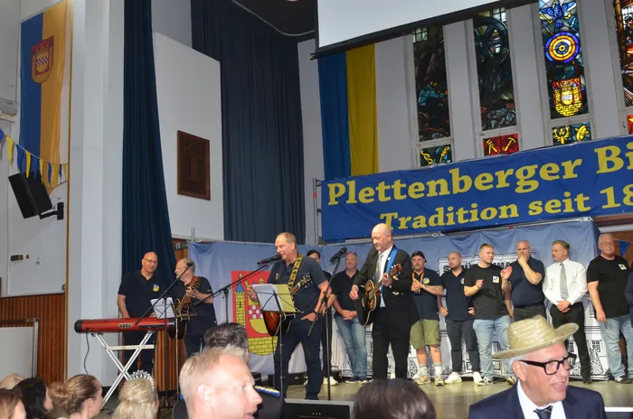 Eine Gruppe von Männern steht auf einer Bühne vor einem Banner mit der Aufschrift „Plettenberger Biergericht Tradition seit 1888“. Einige spielen Instrumente, andere singen oder klatschen. Im Hintergrund sind Buntglasfenster und Wimpelketten zu sehen.