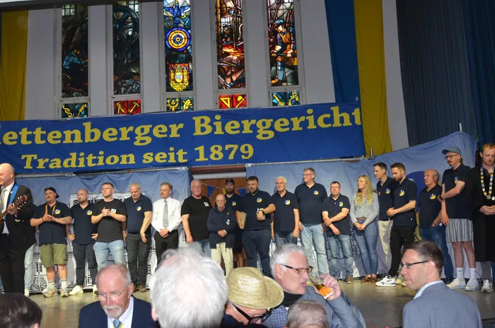Eine Gruppe von Männern und einer Frau steht auf einer Bühne vor einem Banner mit der Aufschrift „Lettenberger Biergericht, Tradition seit 1879“. Im Hintergrund sind Buntglasfenster zu sehen. Im Publikum sitzen weitere Personen.