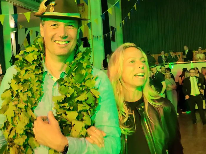 Ein lächelnder Mann mit Hut und einer grünen Girlande steht neben einer lächelnden Frau mit blonden Haaren. Im Hintergrund tanzen Menschen auf einer festlich beleuchteten Tanzfläche.