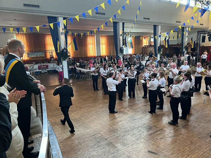 Ein Musikkorps spielt in einer großen Halle mit Parkettboden. Im Hintergrund sind blau-gelbe Wimpelketten zu sehen, und Menschen stehen und sitzen um die Musiker herum.