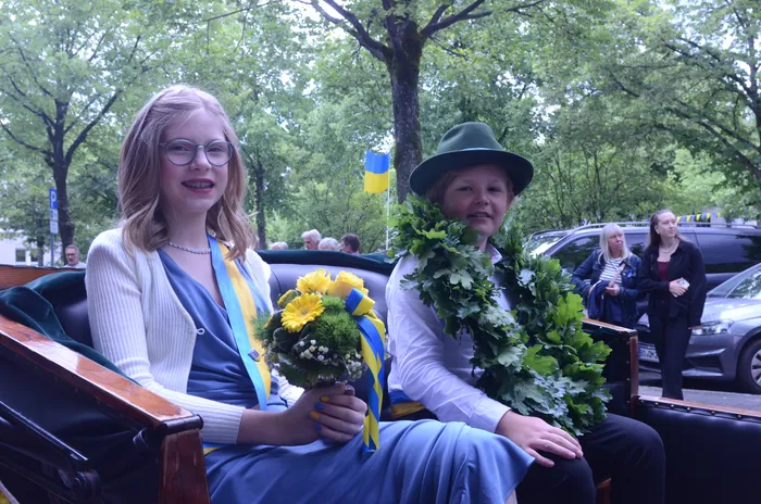 Zwei junge Frauen sitzen in einer mit Holz verkleideten Kutsche. Sie tragen Girlanden und halten Blumensträuße in den Farben Blau und Gelb, während im Hintergrund eine kleine ukrainische Flagge zu sehen ist.
