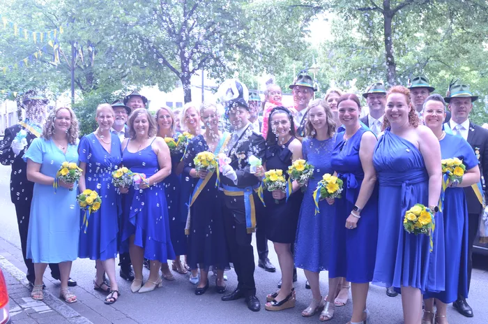 Eine Gruppe von Menschen, darunter Frauen in blauen Kleidern und Männer in traditionellen Uniformen, steht lächelnd für ein Foto zusammen. Gelbe Blumensträuße werden gehalten, während Seifenblasen um sie herum schweben.