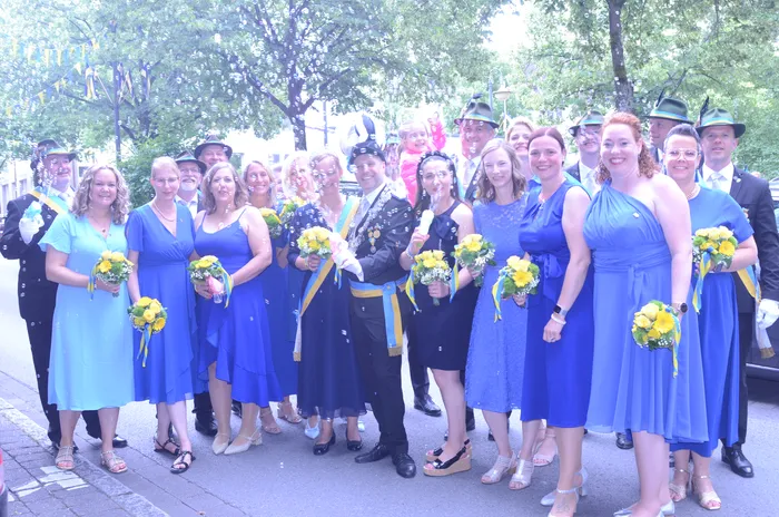 Eine Gruppe von etwa 15 Personen, hauptsächlich Frauen, steht lächelnd in einer Reihe. Die meisten tragen blaue Kleider und halten gelbe Blumensträuße. Ein Mann in einer dunklen Uniform mit vielen Orden steht in der Mitte der Gruppe. Im Hintergrund sind weitere Personen und ein Festwagen zu sehen.