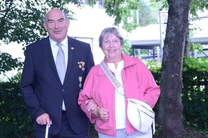 Ein älterer Mann in Anzug mit Orden und eine ältere Frau mit Jacke stehen lächelnd nebeneinander. Im Hintergrund sind Bäume und Gebäude zu sehen.