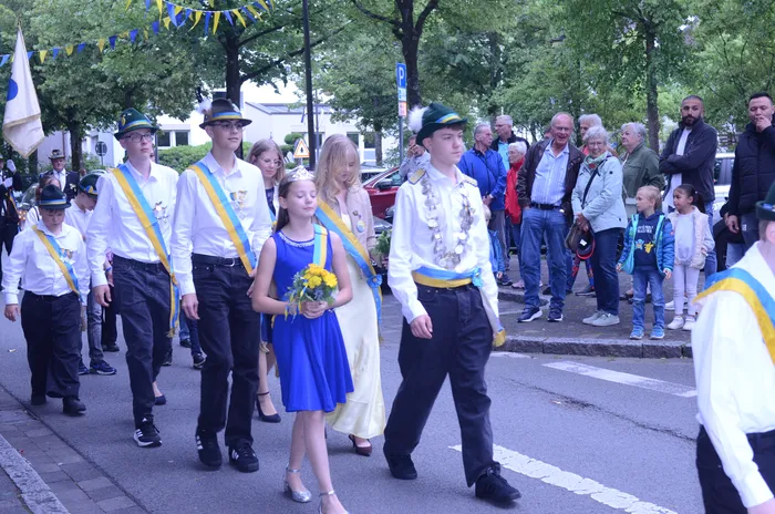 Eine Gruppe von Jugendlichen und Erwachsenen marschiert in Uniformen und mit Orden durch eine Stadt. Vorne trägt ein Mädchen ein blaues Kleid und eine Krone, während sie einen Blumenstrauß hält. Im Hintergrund stehen Zuschauer am Straßenrand.